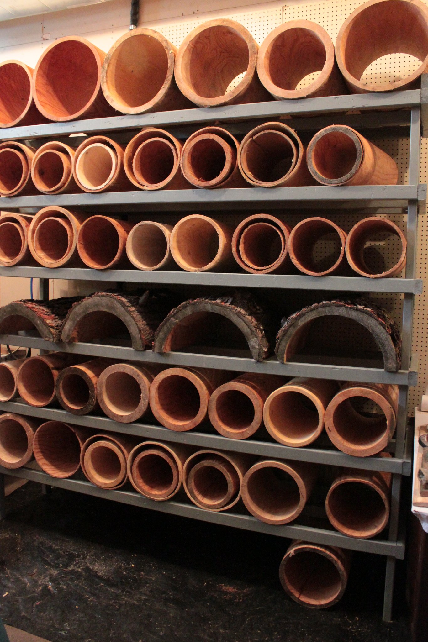 Floor-to-ceiling steel shelving rack packed with finished IC WOOD hollow log cylinders and half-log pieces — warm red pine grain, production shop interior, March 2014